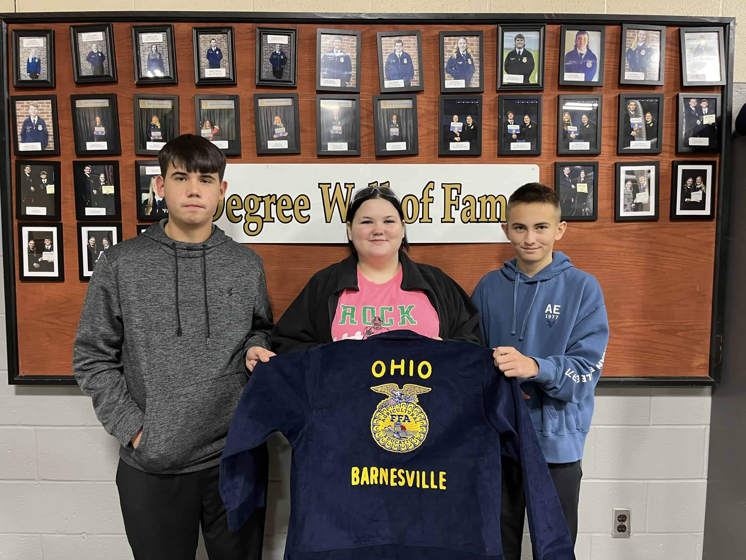 Three Barnesville FFA Freshman–Caleb Bush, Chloe Gordon, and Aiden Starr–stand in front of the Degree Hall of Fame holding their new blue FFA jacket embroidered with "Ohio" and "Barnesville."